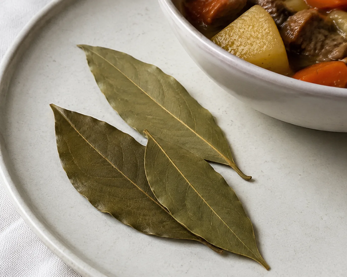 Three whole dried bay leaves on a pale plate beside a bowl of stew, stiff and completely intact with their wavy edges unbroken