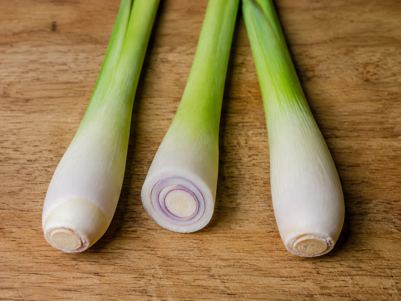 Three cut lemongrass stalks on a wooden board showing the swollen pale white base of each, with one sliced across to show the tight pale rings inside