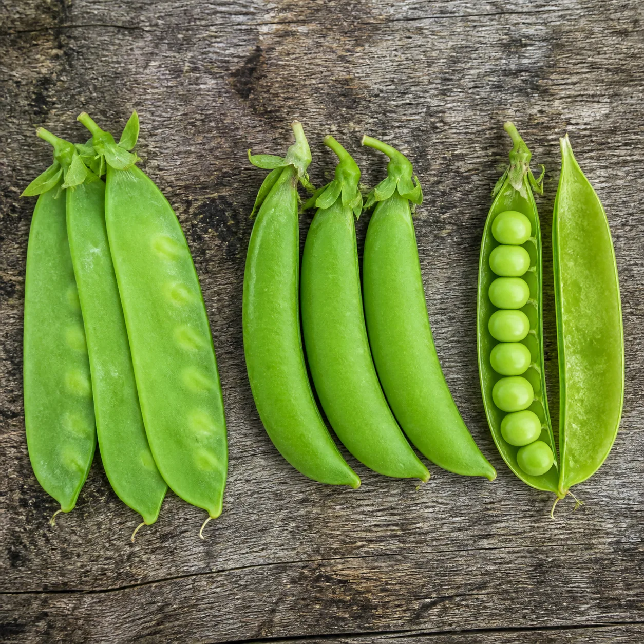 Flat snow pea pods, plump snap pea pods, and a split shelling pea pod showing round peas inside, laid side by side on a wooden board