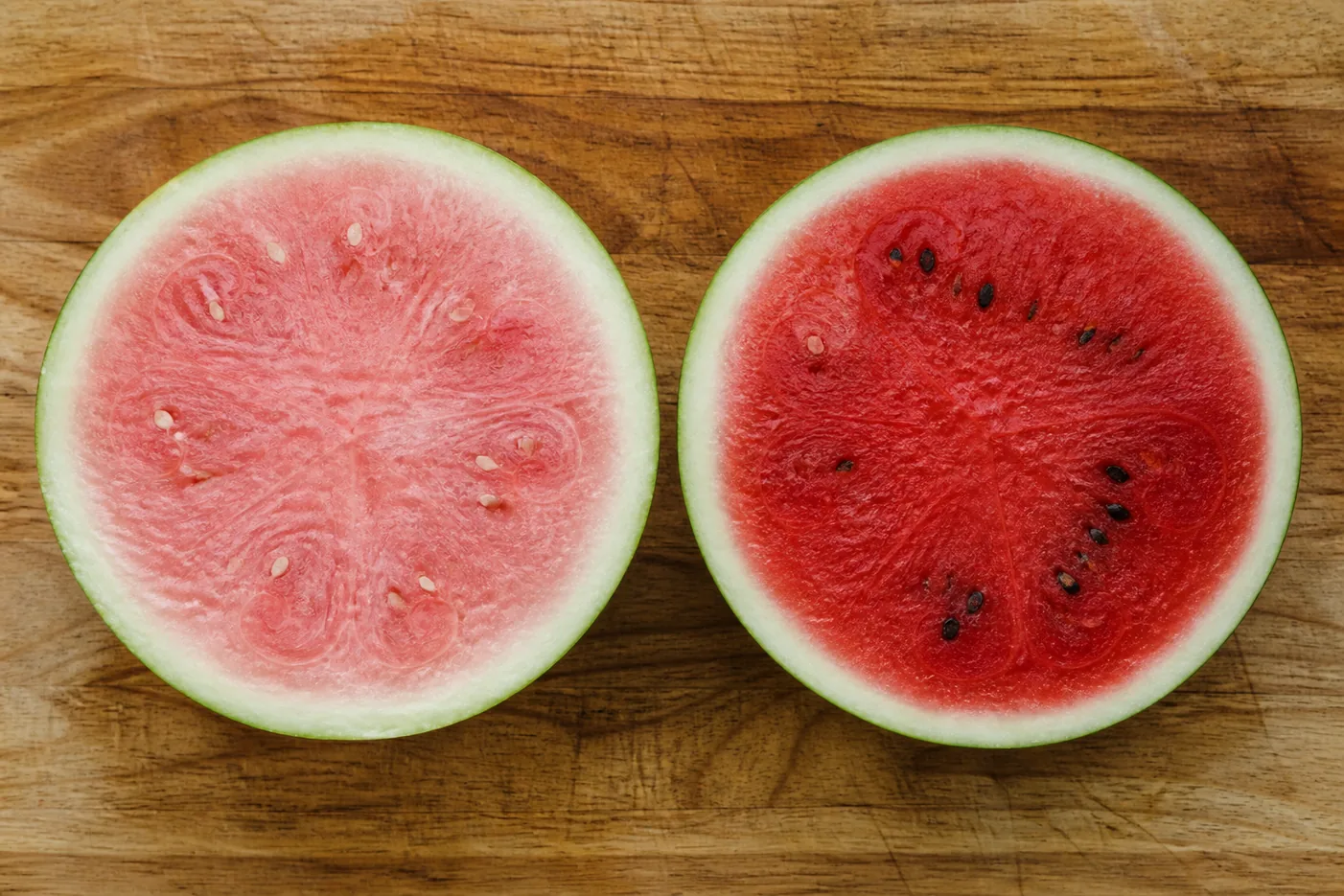 Two watermelon halves cut side by side on a wooden board, the left with pale pink watery under-ripe flesh and white seeds, the right with deep red ripe flesh and dark mature seeds